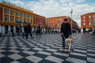 Une personne aveugle accompagnée de son chien guide se promène dans une place de la ville de Nice. On voit au loin des bâtiments colorés qui confèrent à la place un certain charme et au sol des damiers. 
