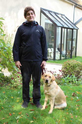 Raphaël se tenant debout, à gauche de Pouxie, son chien guide d'aveugle, labrador sable. ￼