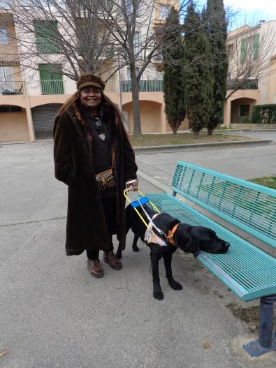 Une femme avec un long manteau noir et un chapeau marron qui tiens son chien guide, un labrador noir qui a un harnais