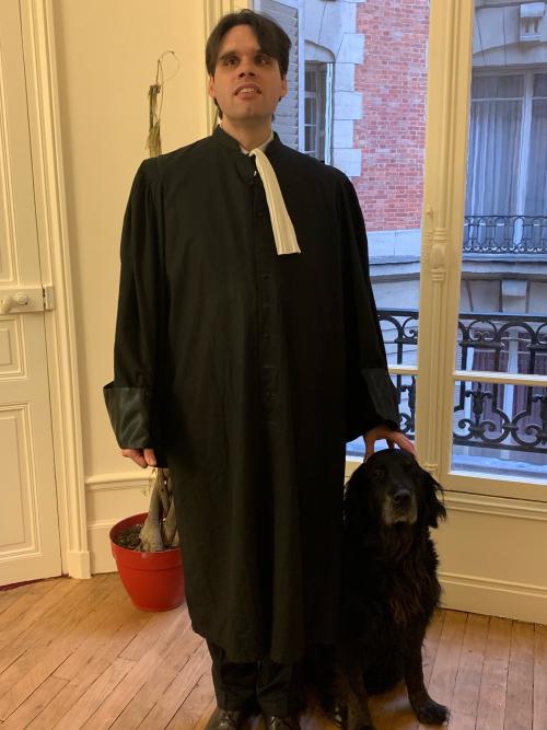 Un homme brun debout avec sa tenue d'avocat. A côté son chien guide. 