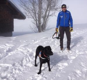 Elie, un homme debout dans la neige avec son chien guide, un labrador noir a ses côtés