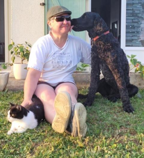 Nathalie assise dans l'herbe, caresse son chat d'un côté et se fait lécher la joue par Sia qu'elle serre affectueusement