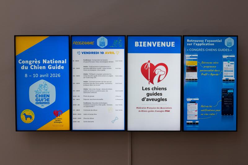 Des écrans d’information numériques, installés à l’entrée du Congrès national des chiens guides 2026, présentent l’identité de l’événement, le programme du jour, l’application accessible et les informations clés pour les participants réunis pendant trois jours à Opio, en France.